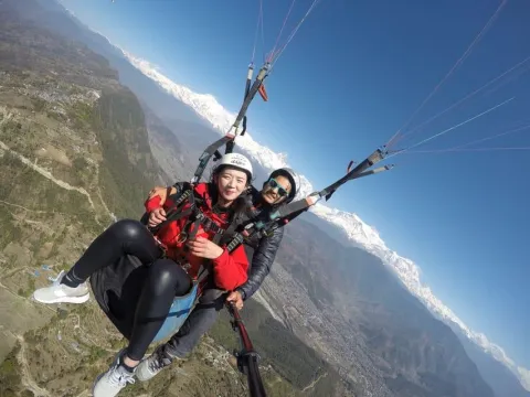 Tandem paraglider soaring above Pokhara with snow-covered Annapurna Himalayan peaks in the background.