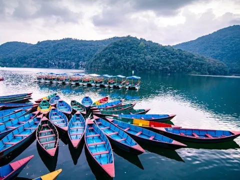 Colorful boats floating on Phewa Lake in Pokhara, Nepal, with green hills and the Annapurna Himalayan range in the background.