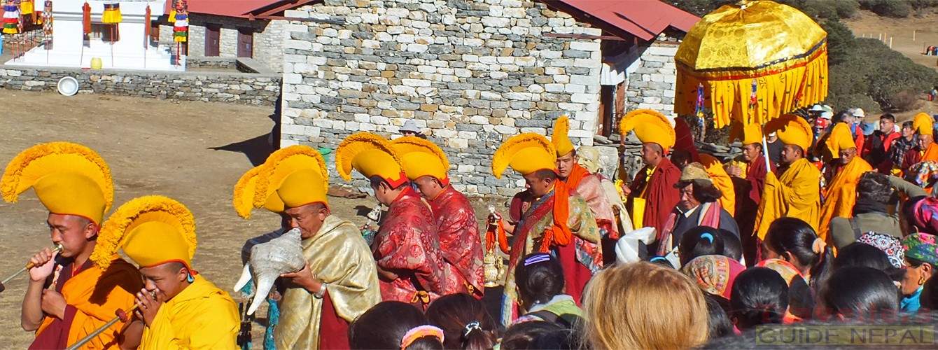 Buddhist monks clad in colorful silk brocade robes and ornate ceremonial masks perform sacred dances in the courtyard of Tengboche Monastery, framed by prayer flags and a snow-capped Himalayan peak under a clear sky.  
