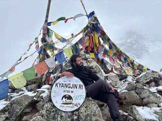 Trekker at Kyanjin Ri viewpoint at 4,700 meters surrounded by colourful prayer flags, snow-covered rocks, and Himalayan mountains.