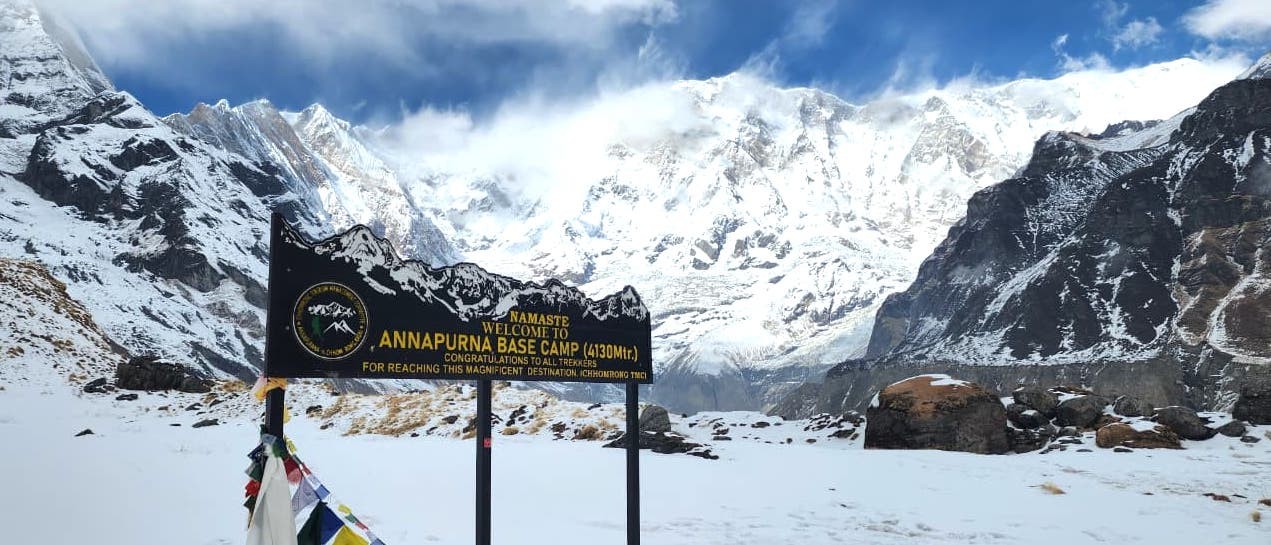 Annapurna Base Camp signboard with Annapurna I towering in the background, surrounded by snow-covered peaks, mountain lodges, and the breathtaking Annapurna Sanctuary.