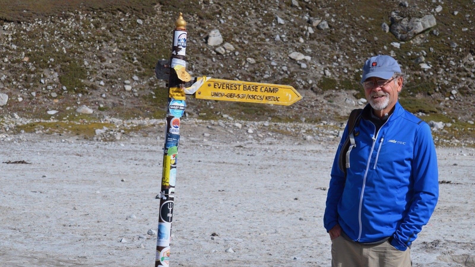 Trekker wearing the three-layer clothing system on the Everest Base Camp Trek in October, including a moisture-wicking base layer, warm fleece mid-layer, waterproof outer shell, hiking pants, boots, gloves, and a backpack against a backdrop of Mount Everest and Ama Dablam. 