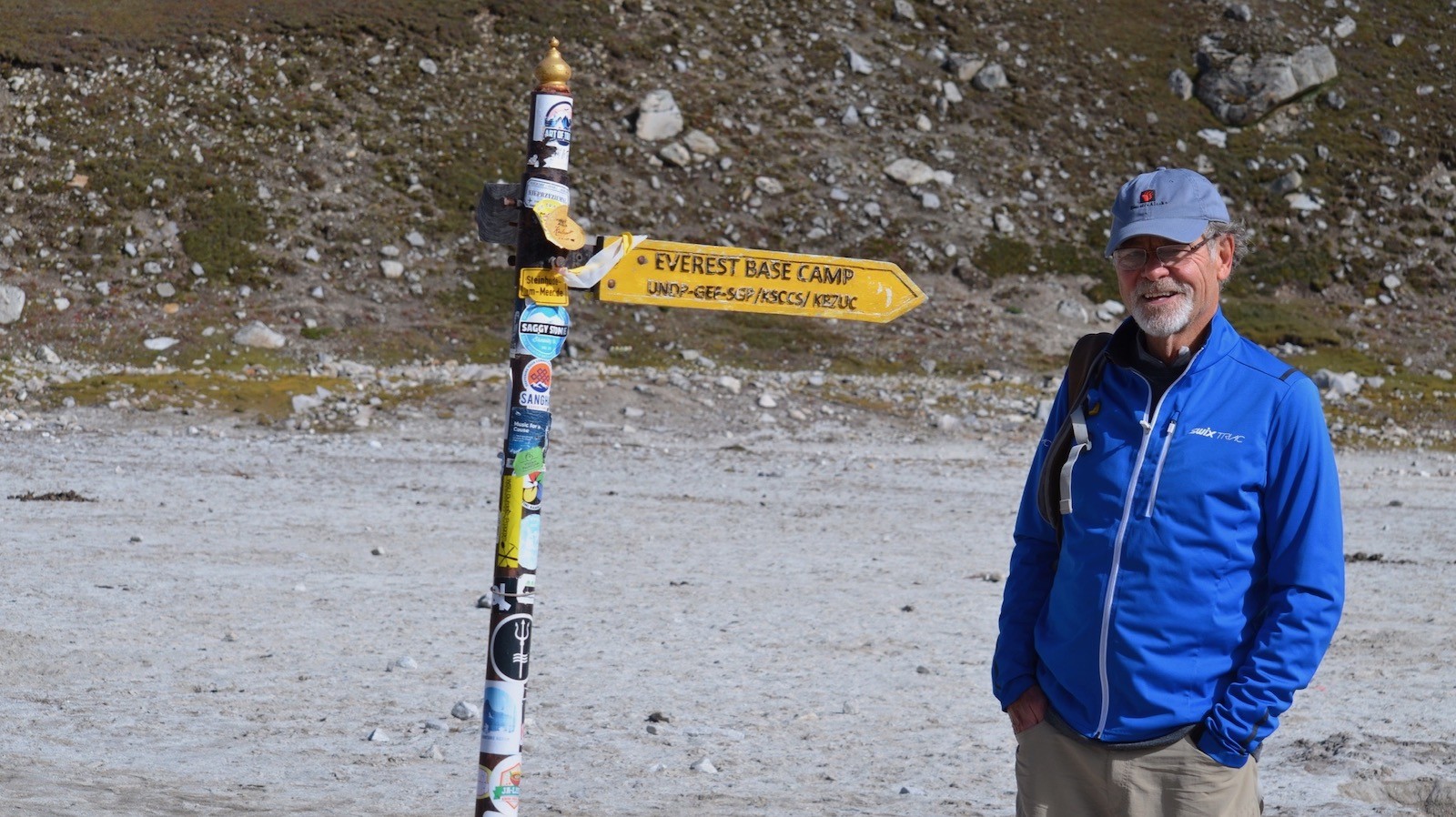 Trekker standing beside the Everest Base Camp trail sign on a rocky alpine landscape during the Everest Base Camp Trek under clear mountain skies.