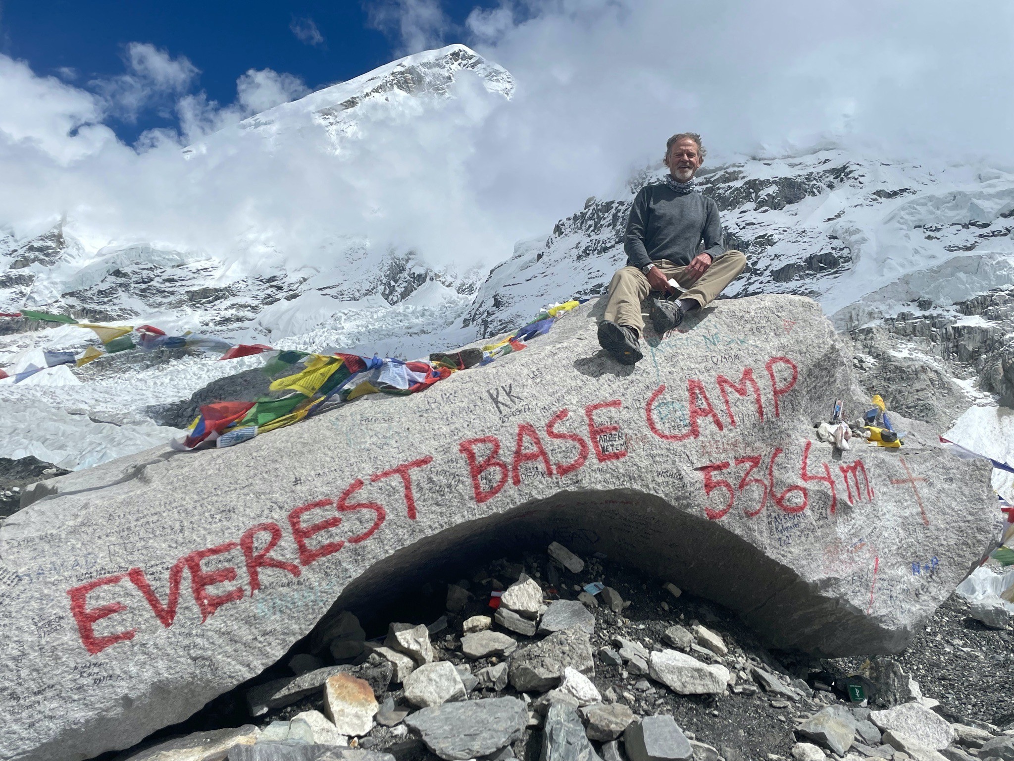 Trekker standing beside the iconic Everest Base Camp rock with colorful prayer flags, the Khumbu Glacier, and towering peaks of Everest and Nuptse under a crystal-clear Himalayan 