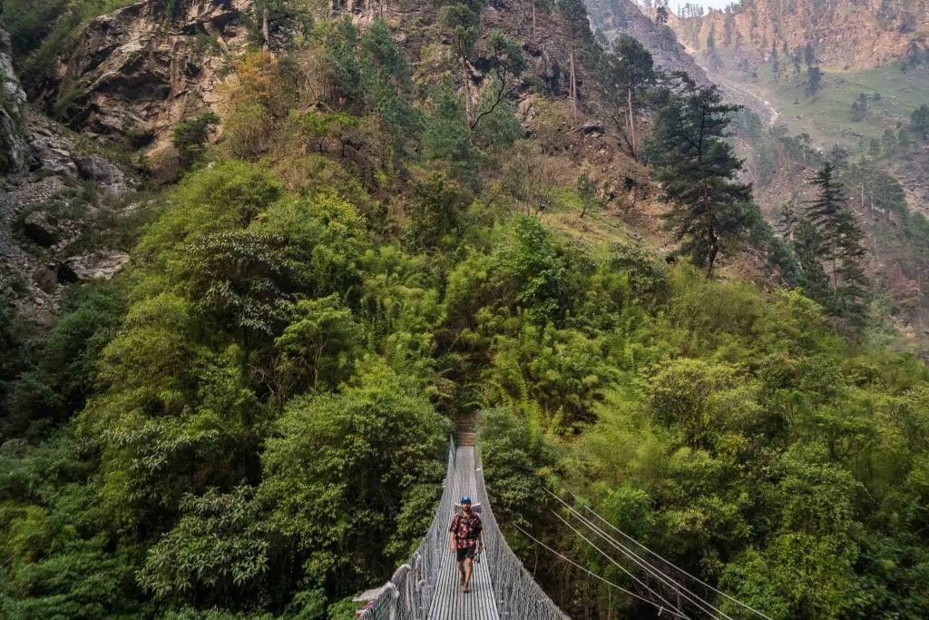 Suspension bridge crossing over a river in the green Tsum Valley, with mountain views and traditional villages in the background.