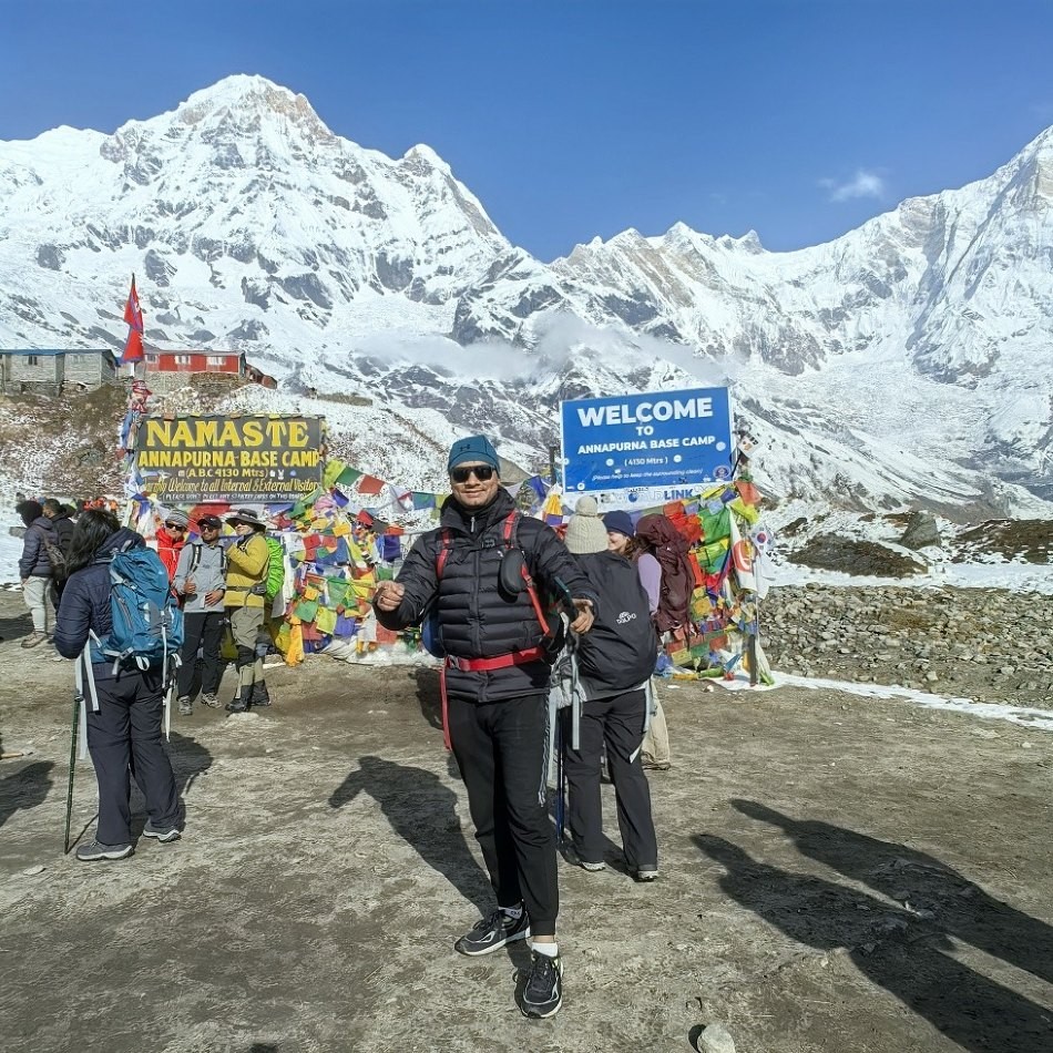 Happy trekker celebrating at Annapurna Base Camp with snow-covered Himalayan peaks and the iconic base camp sign in the background.
