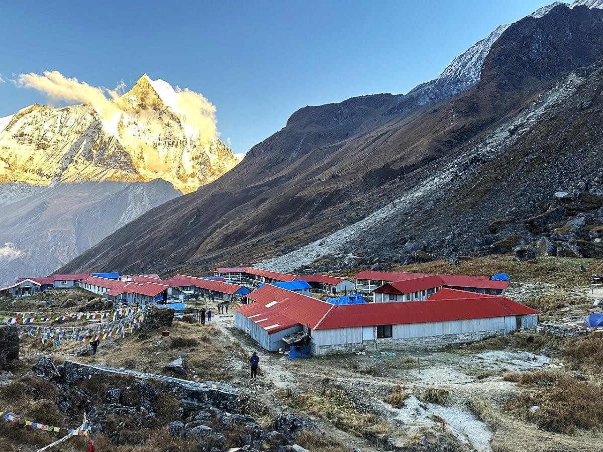 Traditional teahouse lodges at Annapurna Base Camp with Machhapuchhre (Fishtail) towering above, surrounded by the Annapurna Glacier, snow-covered Himalayan peaks, and colorful prayer flags under a clear blue sky.