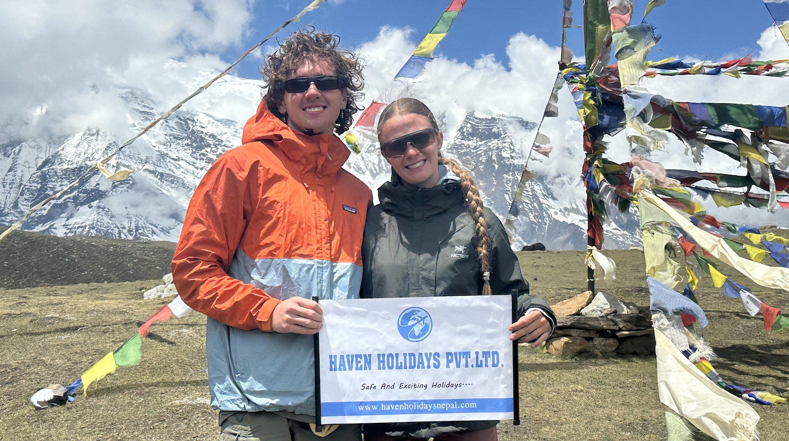 High-altitude alpine trekking gear laid out in Manang, including a down jacket, thermal base layers, fleece, gloves, warm hat, trekking boots, sunglasses, and backpack, with snow-capped Himalayan peaks in the background.