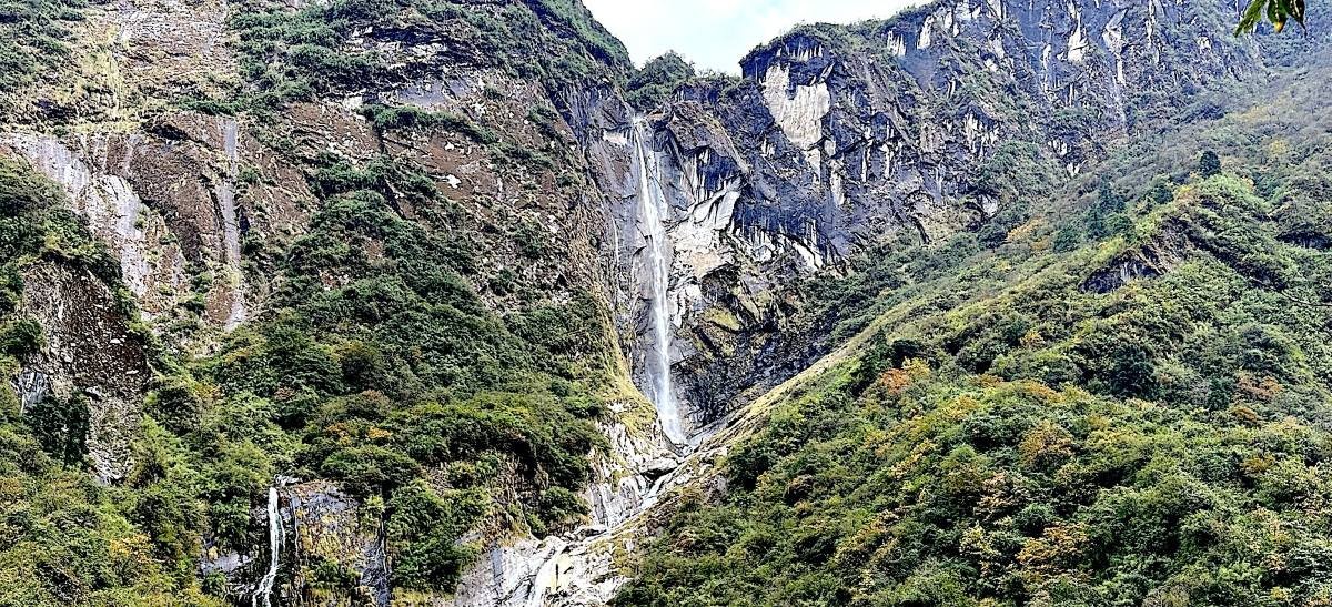 Waterfall cascading through dense Himalayan forest along the Annapurna trekking trail, surrounded by moss-covered rocks, green trees, and misty atmosphere. 