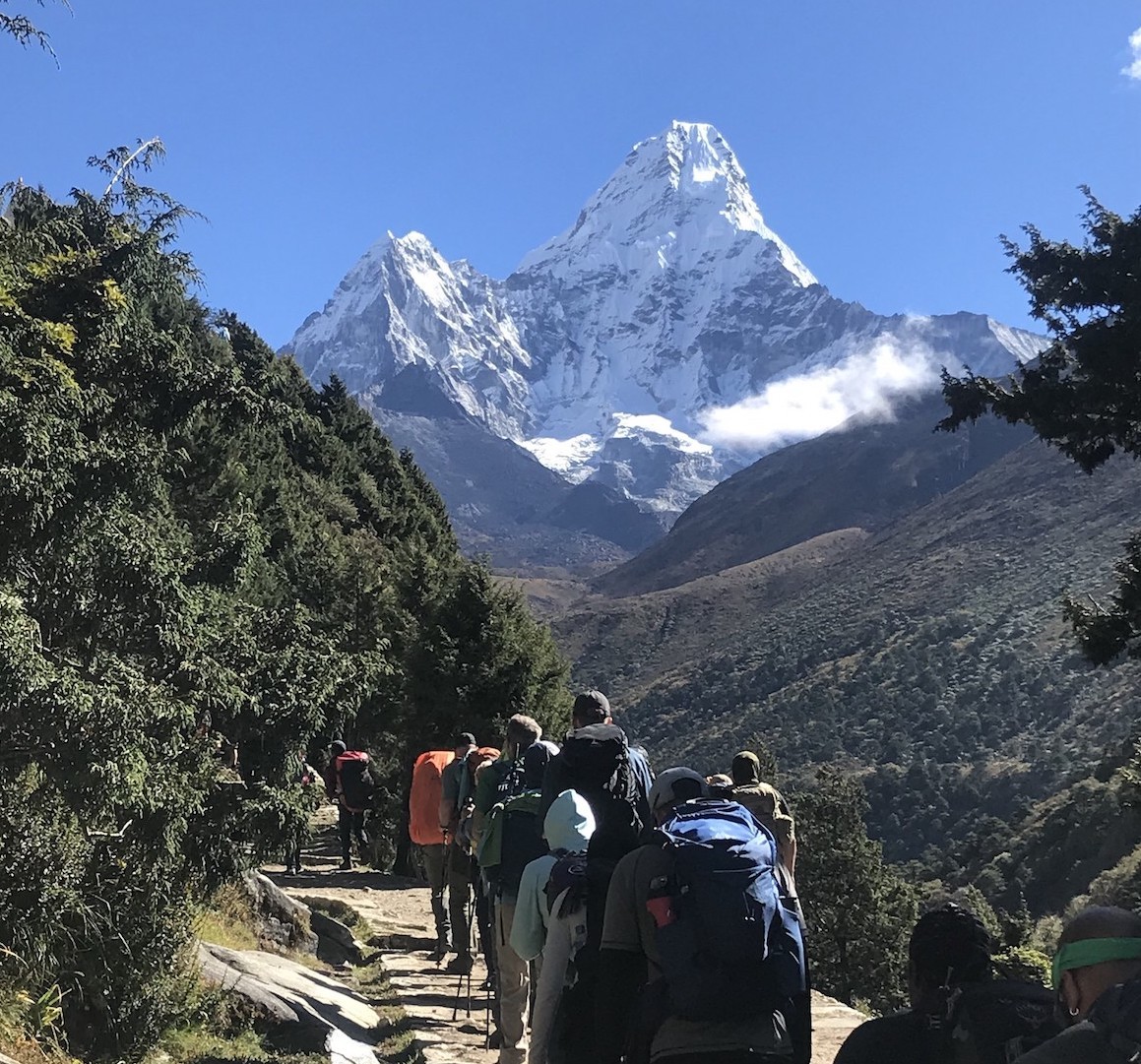 Trekkers walking along the Everest Base Camp trail with the iconic Ama Dablam rising dramatically in the background, surrounded by rugged Himalayan landscapes and clear blue skies.