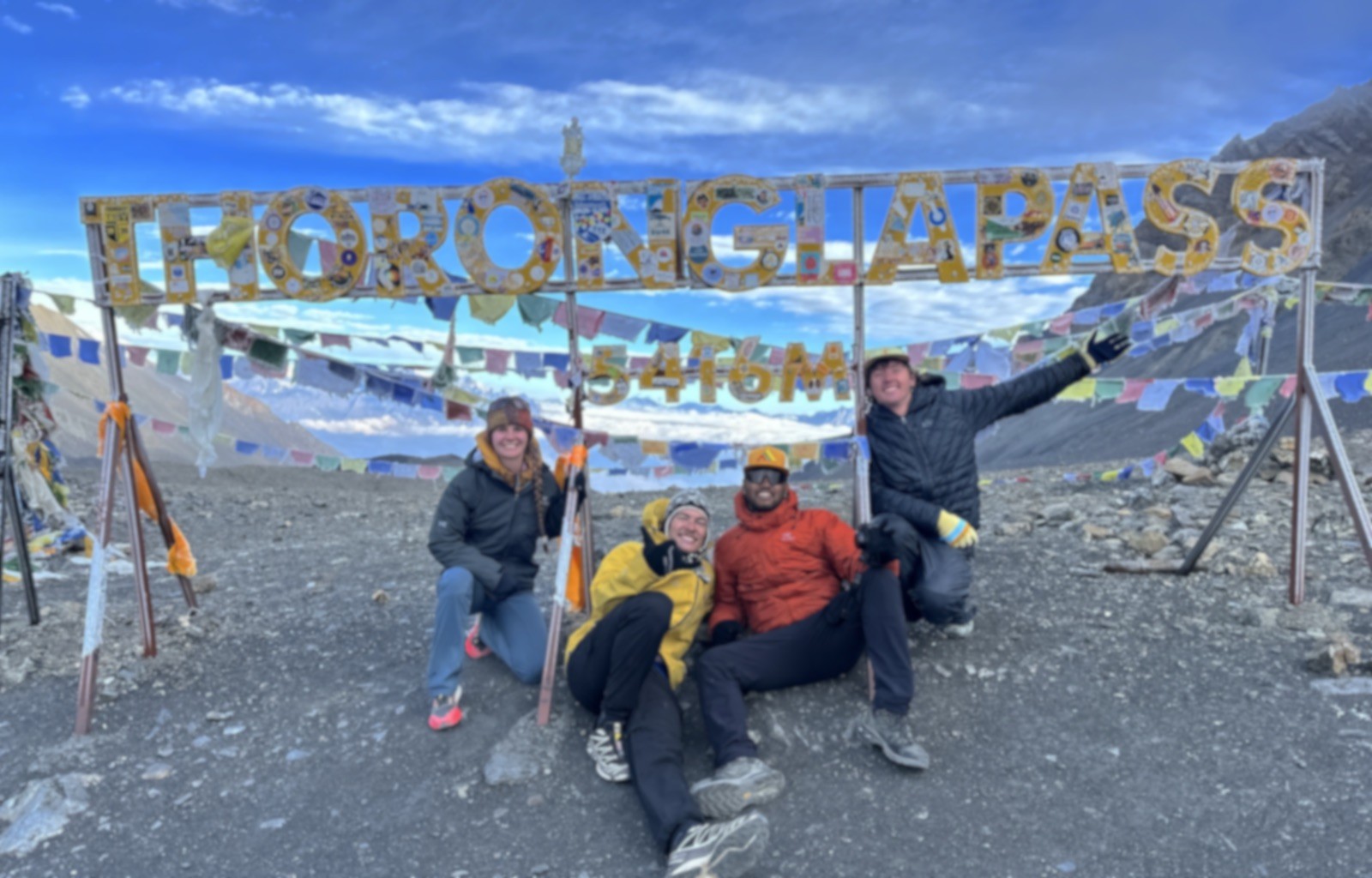 Trekkers crossing Thorong La Pass in September under clear blue skies, with a dry trail, light snow patches, colorful prayer flags, and breathtaking Himalayan views. 