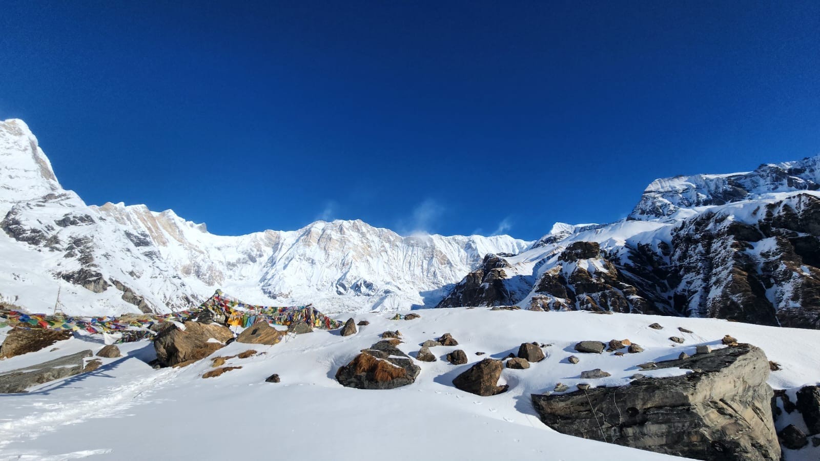 Annapurna Base Camp covered in fresh winter snow, with colorful prayer flags, towering Annapurna I, and the surrounding Himalayan peaks beneath a crystal-clear blue sky in Nepal.