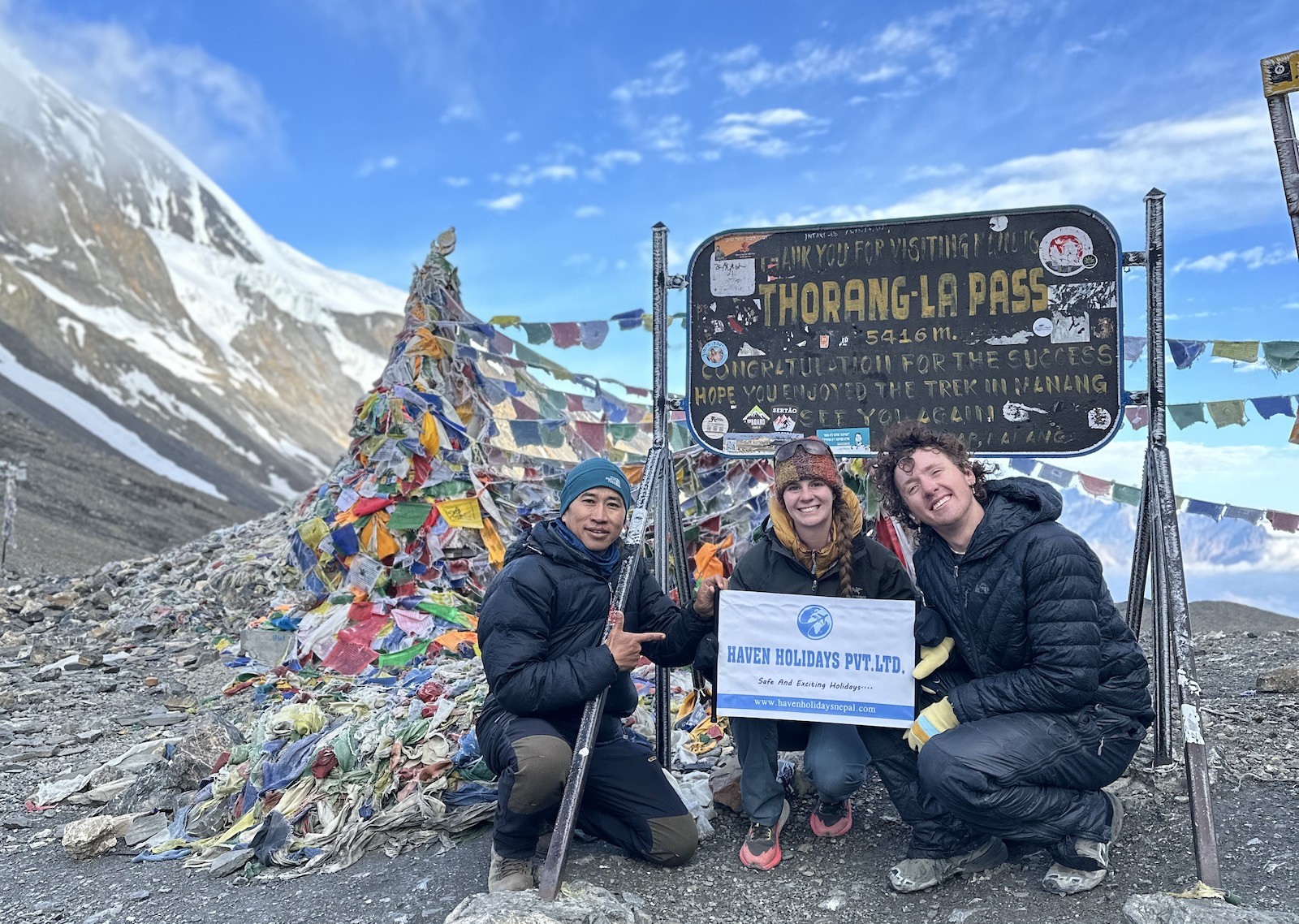 Trekkers crossing Thorong La Pass in October wearing warm layered clothing, insulated jackets, gloves, and hats, with prayer flags, rocky alpine terrain, light snow, and panoramic Himalayan peaks under a clear blue sky.