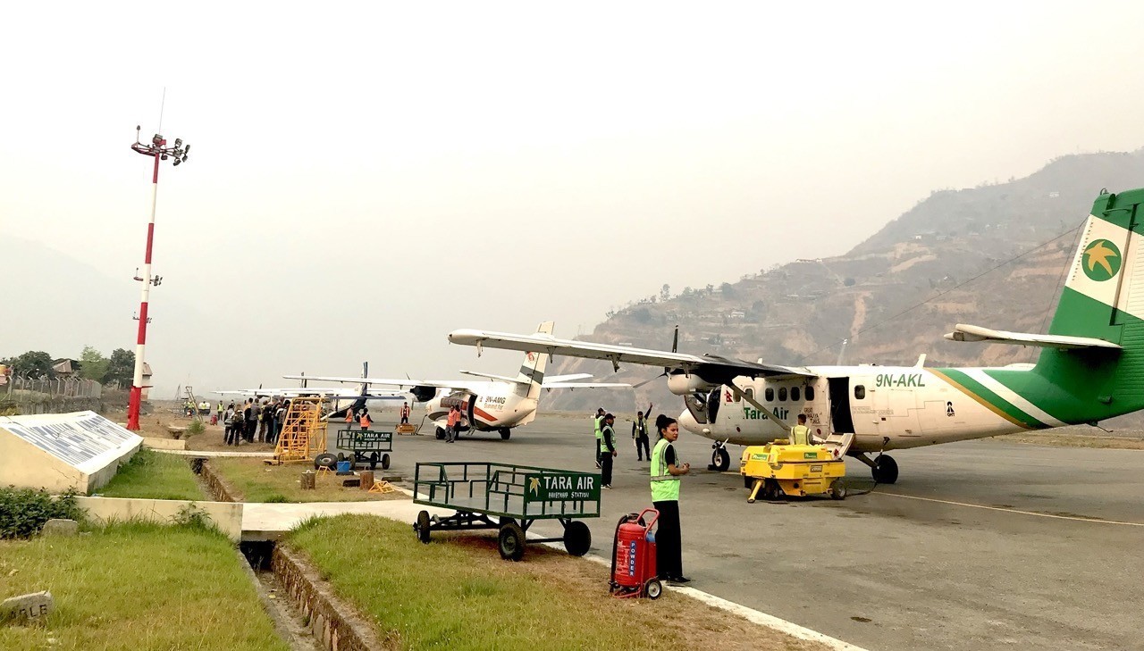 A view of the tarmac at Ramechhap Airport (Manthali Airport) showing several small Twin Otter and Dornier propeller aircraft, including Tara Air planes, parked near airport staff and luggage carts under a hazy, mountain-lined sky.  