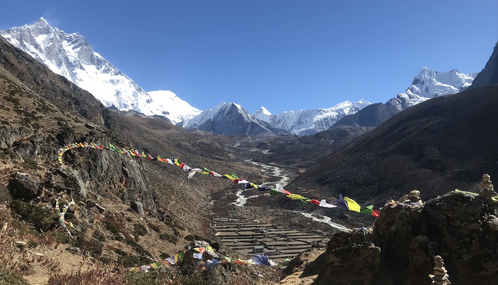 Panoramic view from Dingboche during an acclimatization hike, featuring colourful prayer flags, the Imja Valley, and towering Himalayan peaks under a clear blue sky.