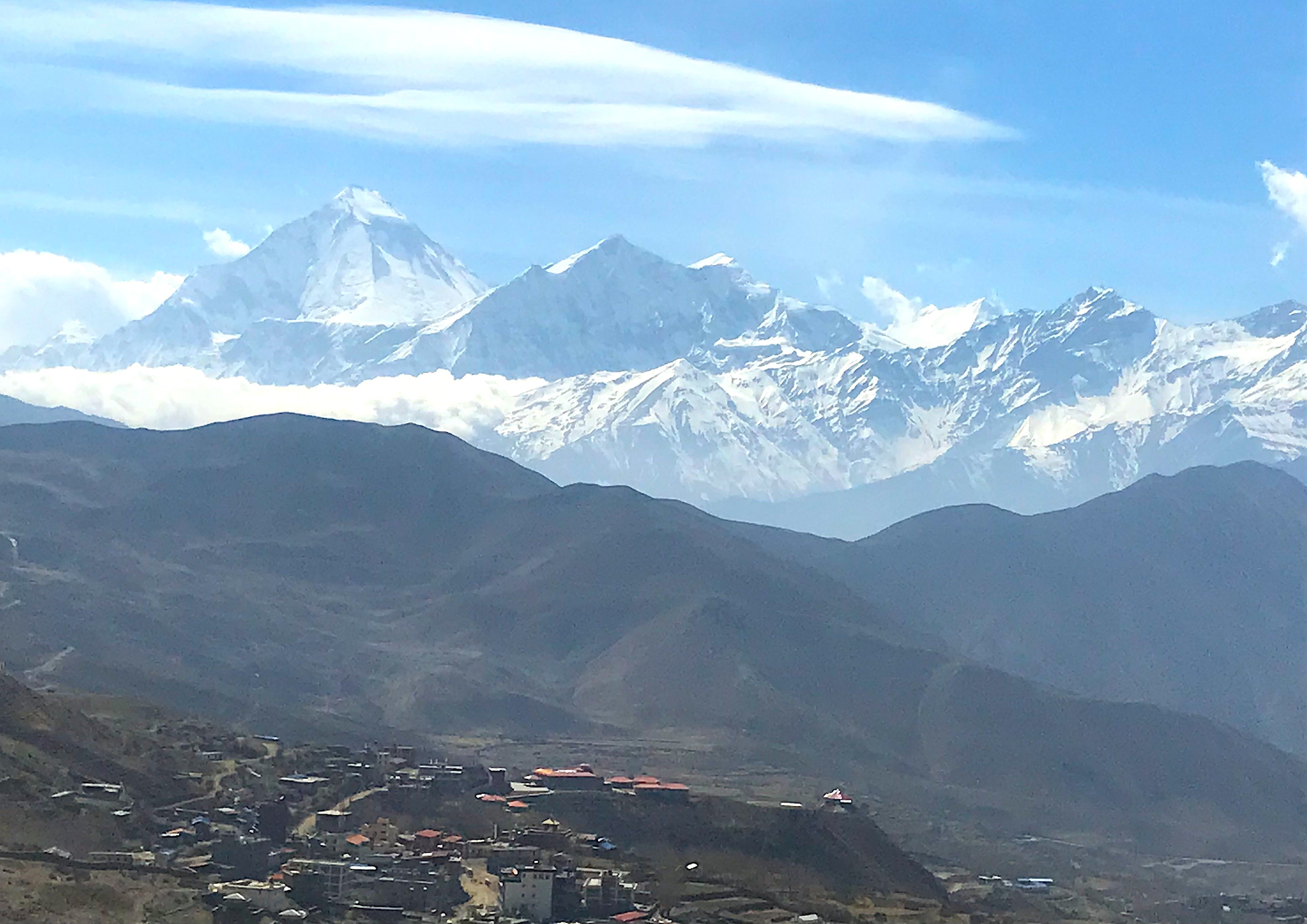 Panoramic view of Mustang with the village below and the snow-covered Dhaulagiri and Annapurna Himalayas rising in the background beneath clear blue skies and lenticular clouds, highlighting Nepal's rain-shadow region in early September.