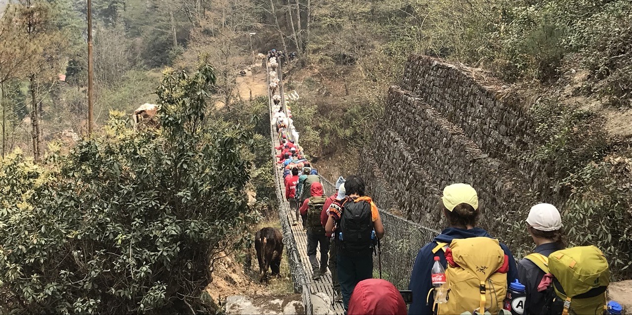 Crowds of trekkers crossing a suspension bridge over a river in the Himalayas, surrounded by lush forested hills and mountain landscapes in Nepal.
