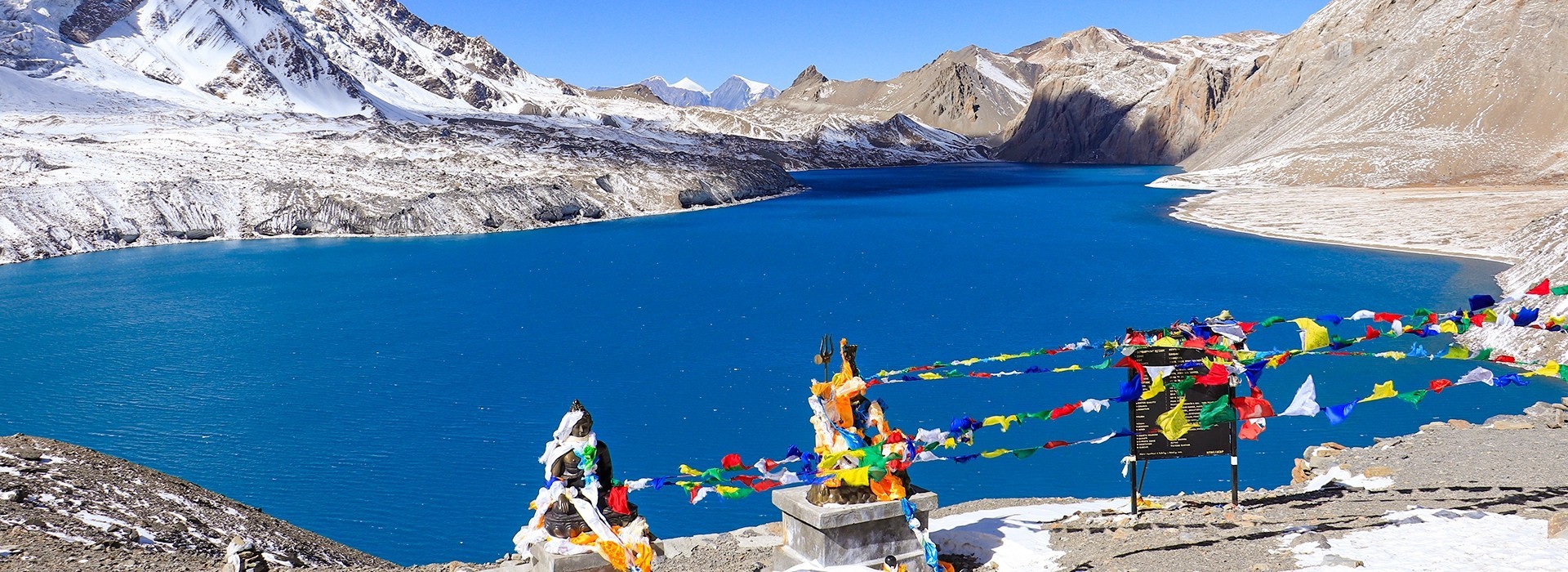Tilicho Lake in October with crystal-clear turquoise water, colorful Buddhist prayer flags, snow-dusted mountains, rocky alpine terrain, and a bright blue sky in the Annapurna region of Nepal.