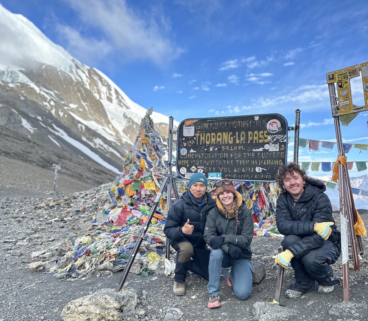 Trekkers crossing Thorong La Pass in cold and windy conditions, wearing proper high-altitude gear including down jackets, gloves, warm hats, insulated pants, and sturdy trekking boots.