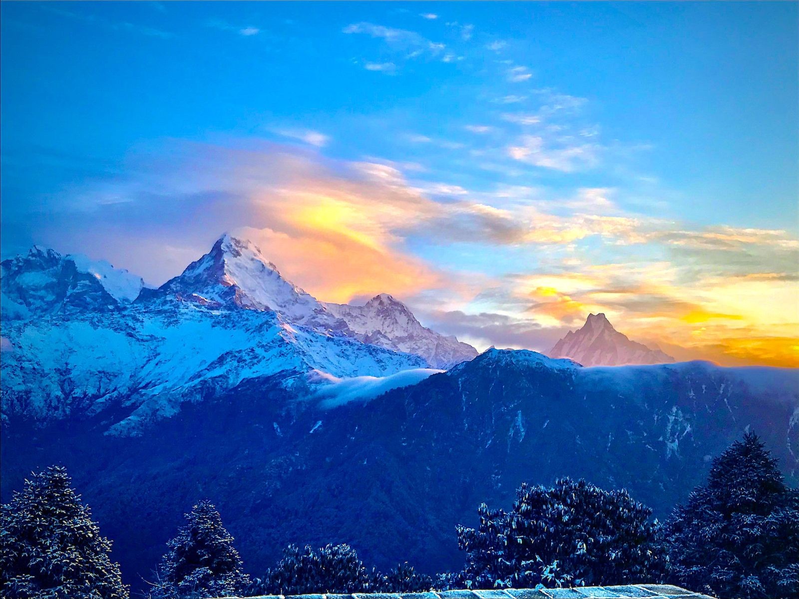 Snow-covered Annapurna South and Machhapuchhre (Fishtail) glowing at sunrise above rhododendron forests, with colorful dawn clouds creating a dramatic Himalayan panorama from Ghorepani Poon Hill.