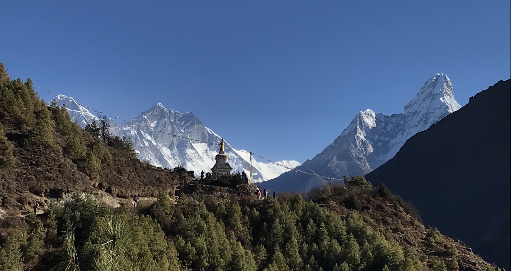 Panoramic view on the trek from Namche Bazaar to Tengboche featuring a Buddhist stupa, Mount Everest, Ama Dablam, and snow-capped Himalayan peaks beneath a clear blue sky.