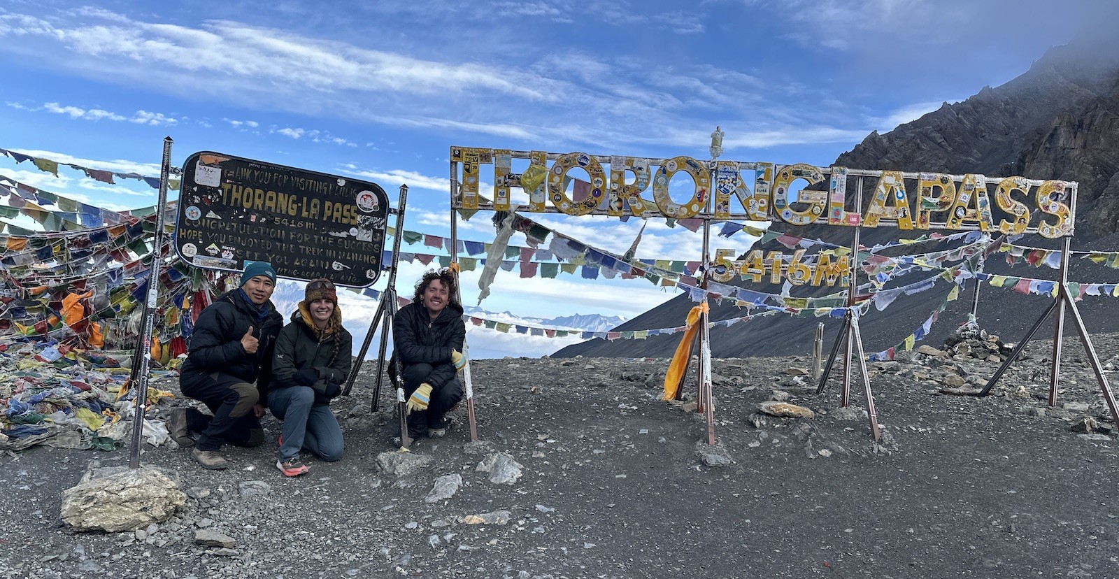 Trekkers celebrating at the summit of Thorong La Pass beside colorful prayer flags and the iconic pass sign, surrounded by snow-covered Himalayan peaks and high-altitude alpine terrain.