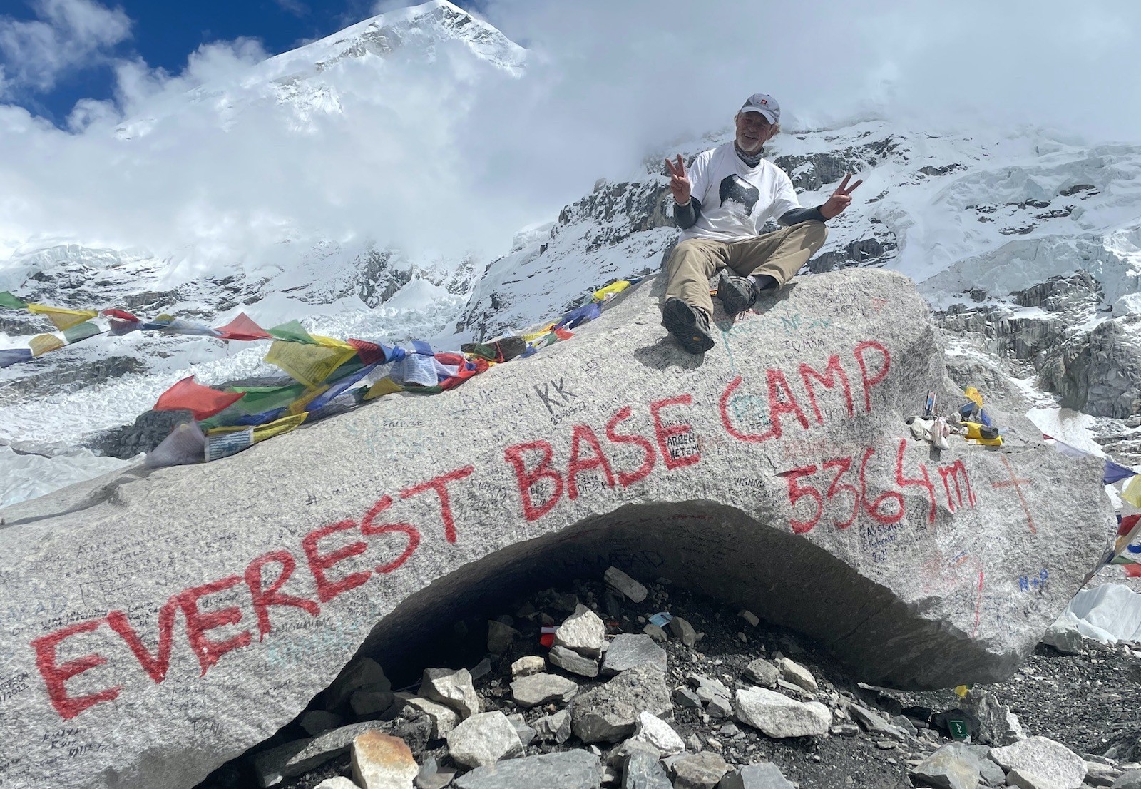 Trekker sitting on the iconic Everest Base Camp marker at 5,364 m, surrounded by colorful prayer flags, glaciers, and snow-covered Himalayan peaks.