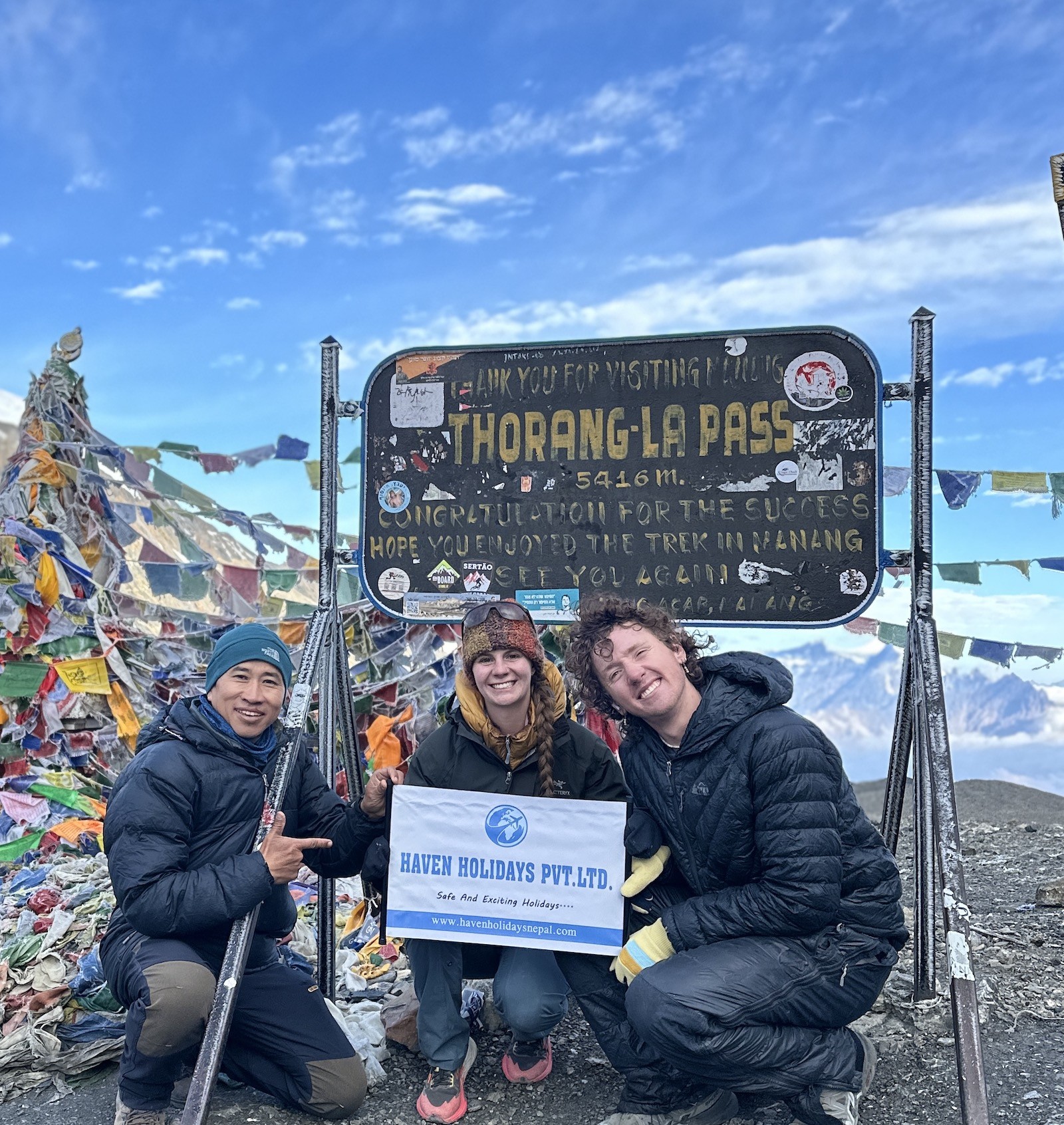 Trekkers crossing Thorong La Pass surrounded by snow-covered Himalayan terrain, prayer flags, and high-altitude mountain scenery.