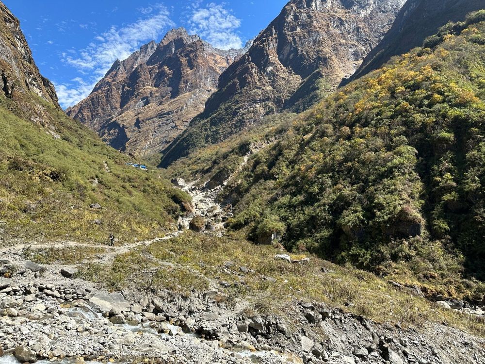 A trekker named George walking on the Annapurna Base Camp Trek trail, heading toward Deurali through a narrow Himalayan valley surrounded by steep cliffs, forested slopes, and snow-covered peaks in the distance.