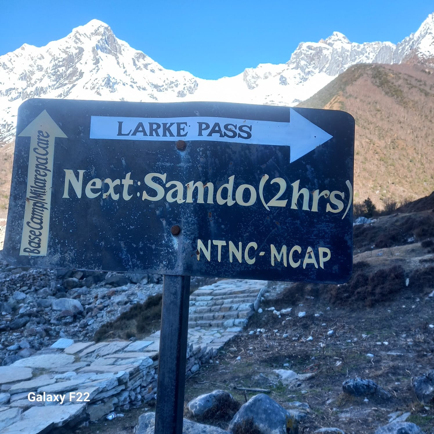 Trail signboard in the Himalayas with snow-capped mountain peaks in the background along a trekking route in Nepal.