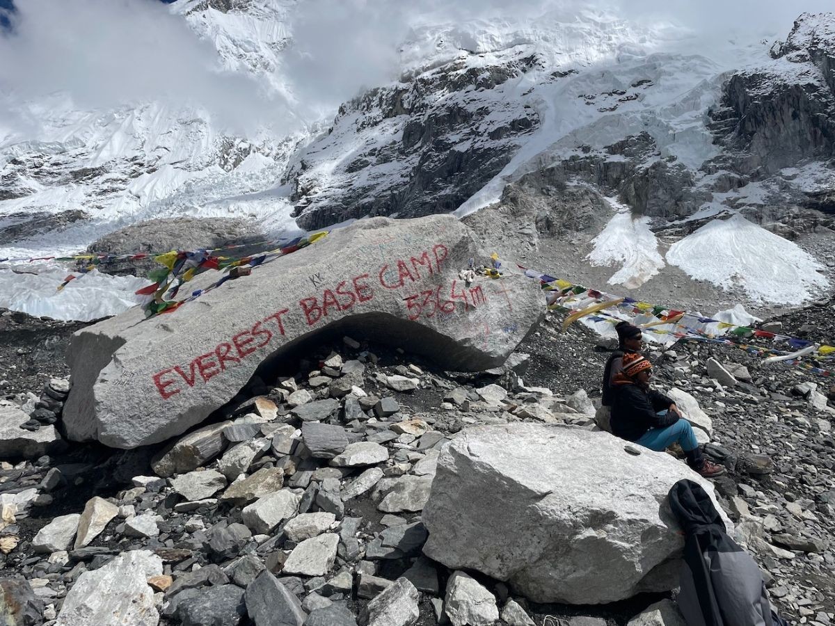 "Trekker standing beside the iconic Everest Base Camp rock with colorful prayer flags, the Khumbu Glacier, and snow-covered Himalayan peaks in the background" Photo