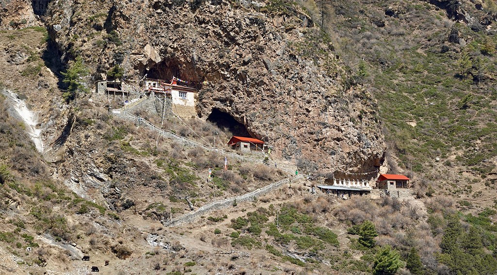 Milarepa Cave above Manang surrounded by rugged cliffs, colorful prayer flags, alpine terrain, and panoramic views of the Annapurna Himalayas, reached via a scenic acclimatization hike on the Annapurna Circuit Trek.
