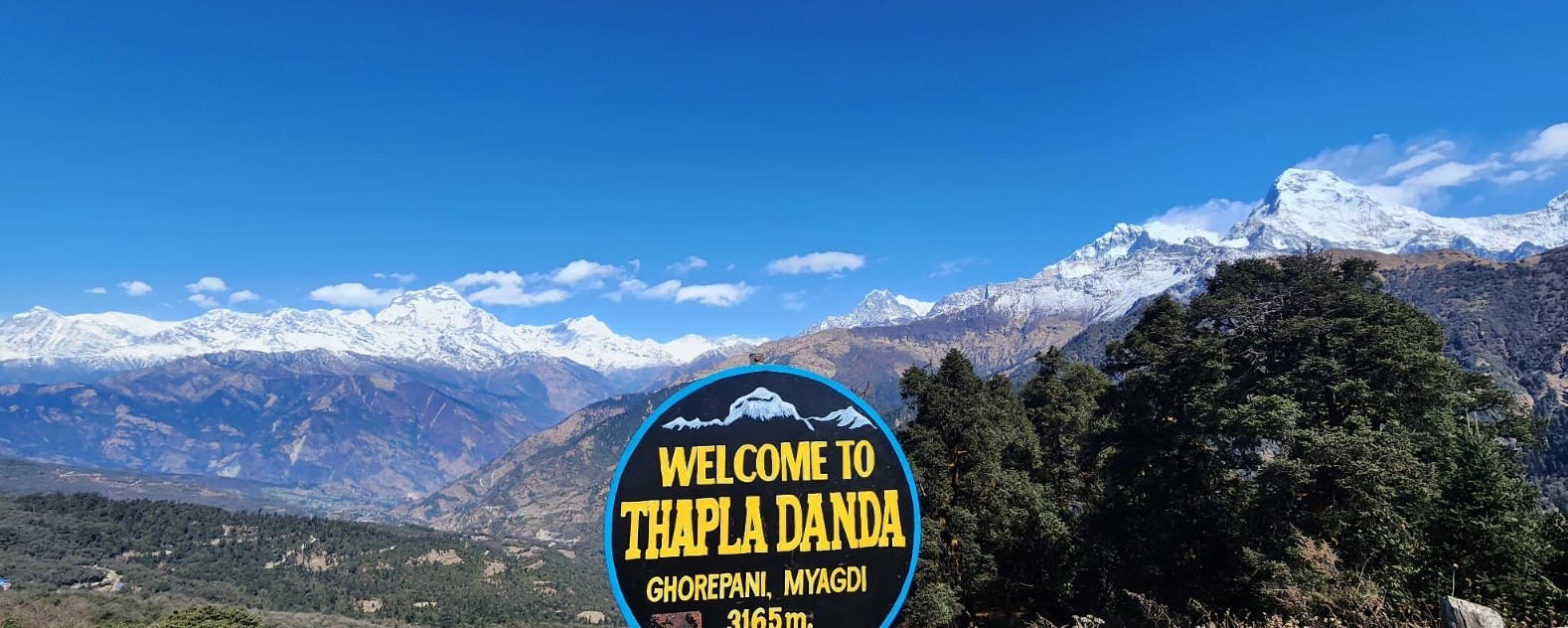 Welcome to Thapla Danda signboard near Ghorepani with panoramic views of Dhaulagiri, Annapurna South, Hiunchuli, and the snow-capped Himalayas under a clear blue sky.