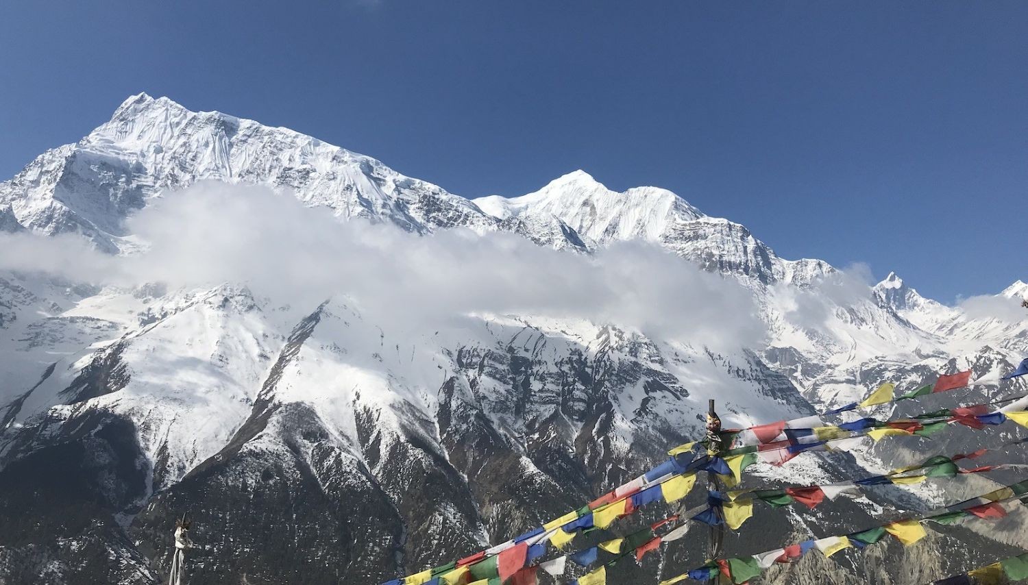 Colorful Tibetan prayer flags flutter in the foreground as snow-covered Annapurna peaks rise above drifting clouds under a deep blue September sky on the Annapurna Circuit Trek.