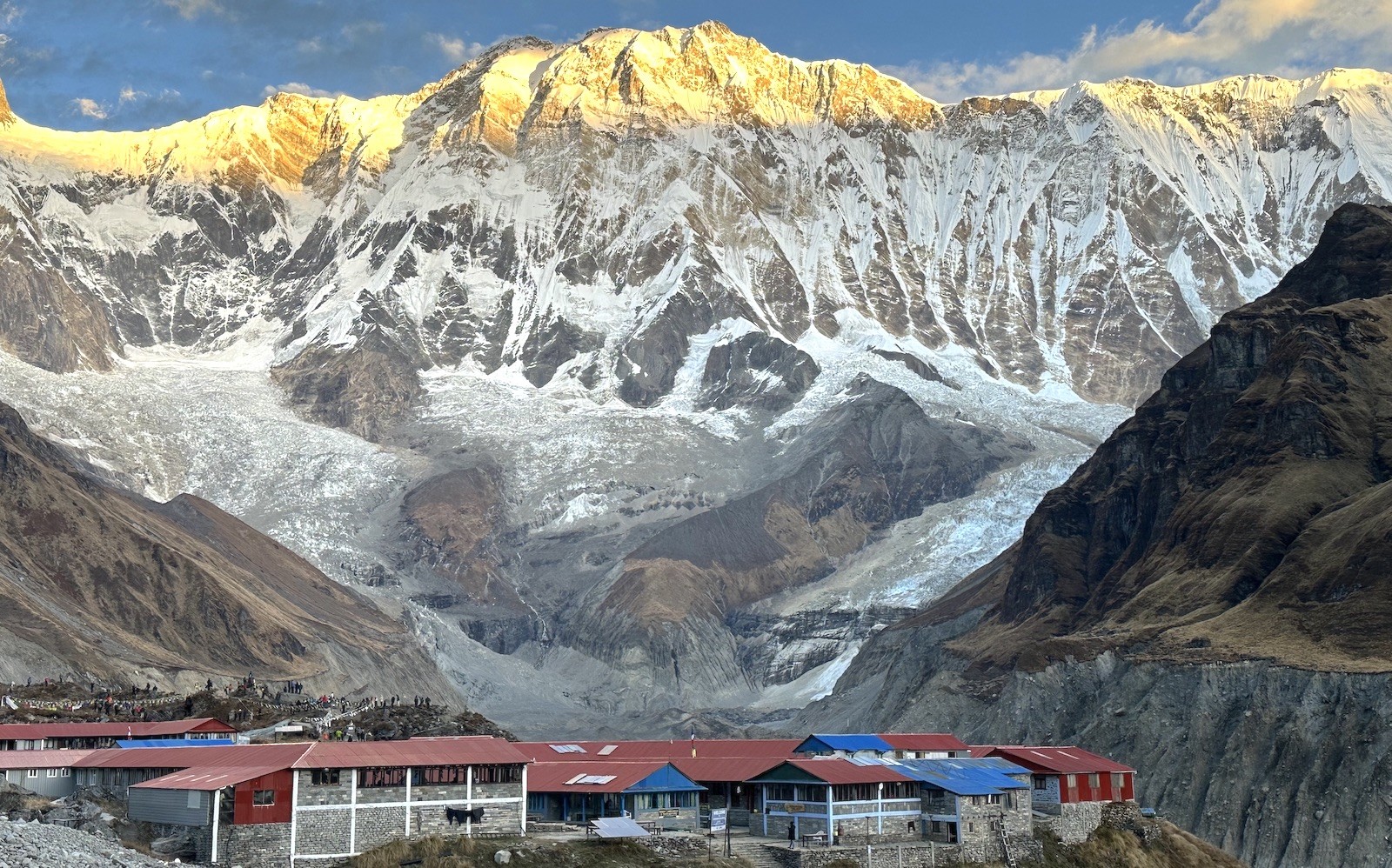 Annapurna Base Camp lodges beneath the towering Annapurna I peak and glacier, illuminated by golden sunrise in the Annapurna Sanctuary.