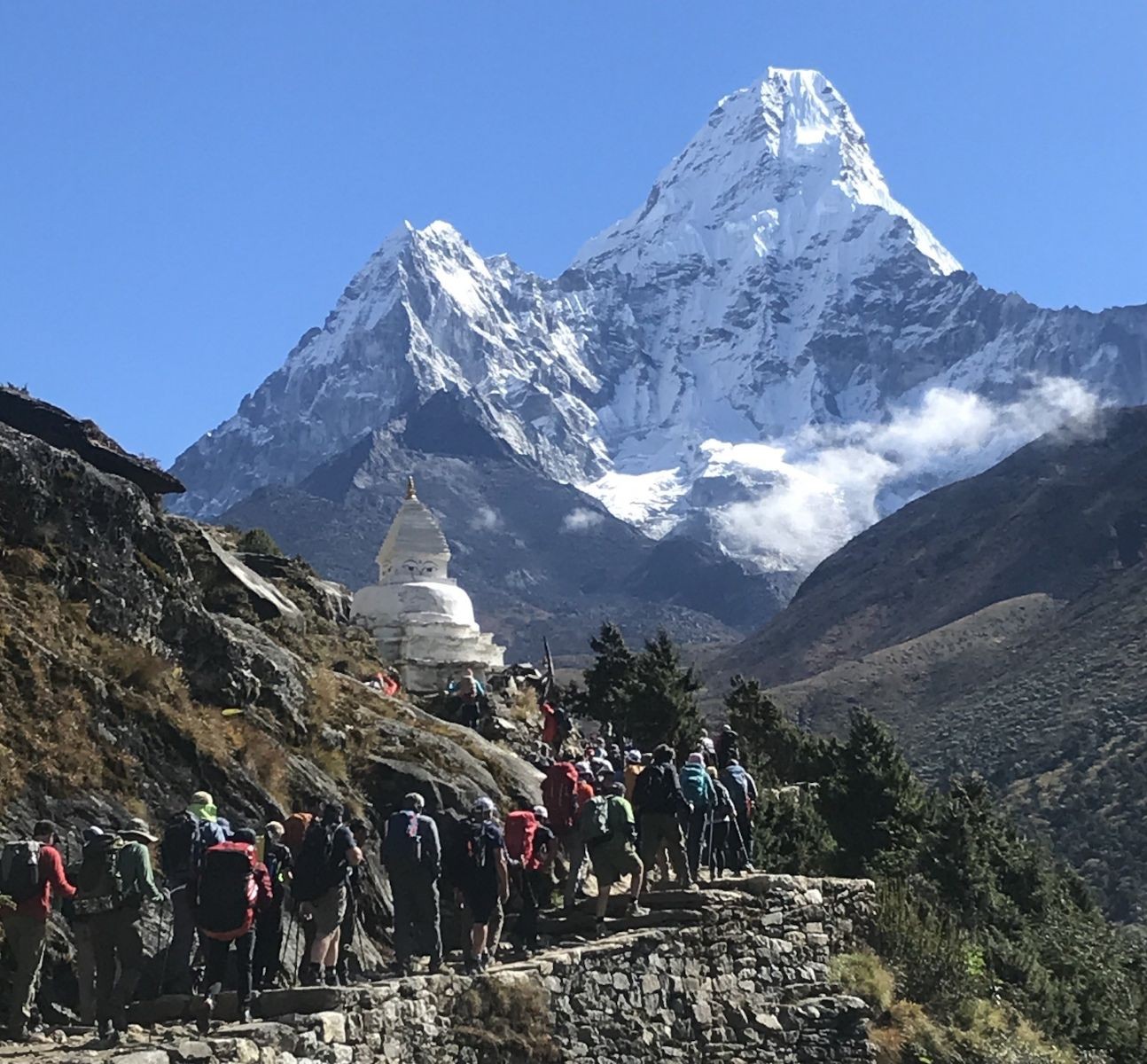 A large group of trekkers with backpacks hikes along a stone trail past a white Buddhist stupa, heading toward the towering, snow-covered peak of Ama Dablam under a clear blue October sky.  