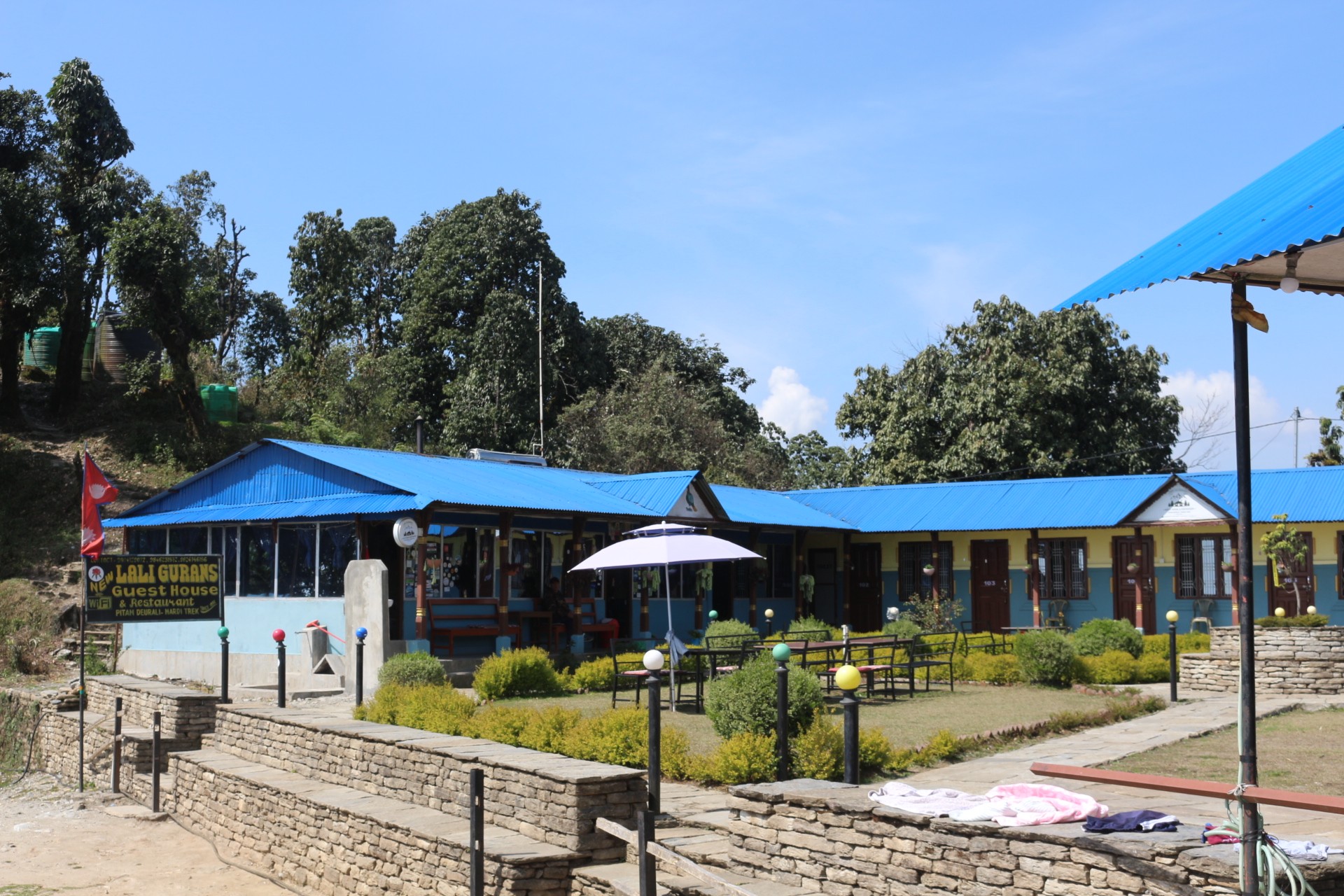 Comfortable teahouse lodge with blue-roofed buildings, private rooms, a landscaped garden, and outdoor seating at Pitam Deurali on the Mardi Himal Trek, surrounded by lush forest under a clear blue sky.  