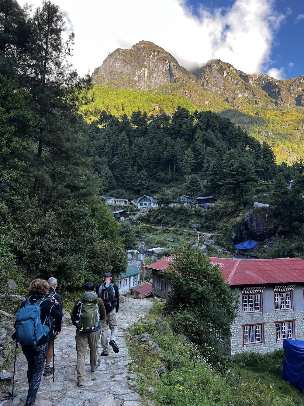 Trekkers walking through a traditional Sherpa village surrounded by lush green pine forests and towering Himalayan mountains during Nepal's monsoon trekking season.