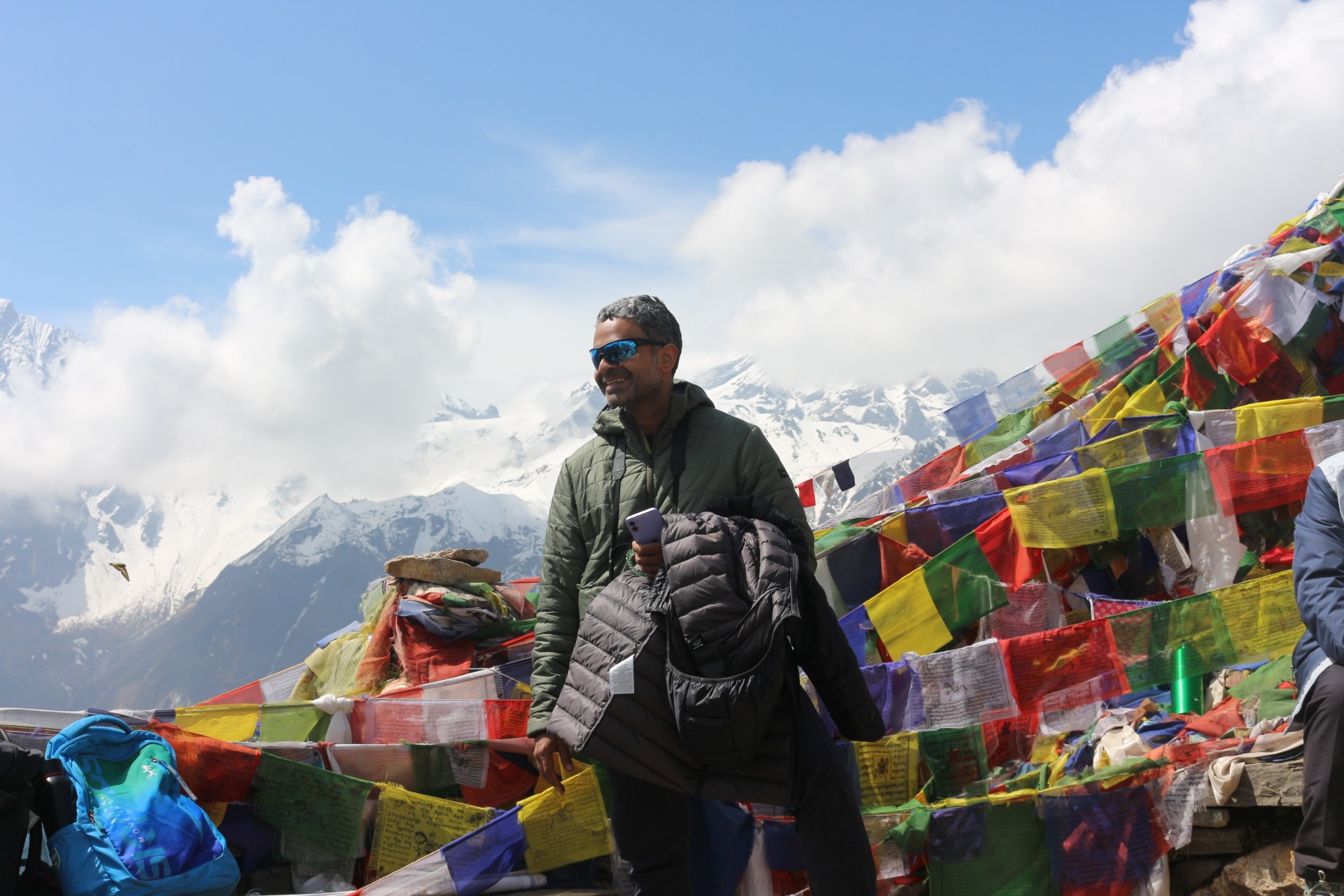 Trekker standing among colorful Buddhist prayer flags with snow-capped Langtang Himalayan peaks and drifting clouds during the Langtang Valley Trek in September.