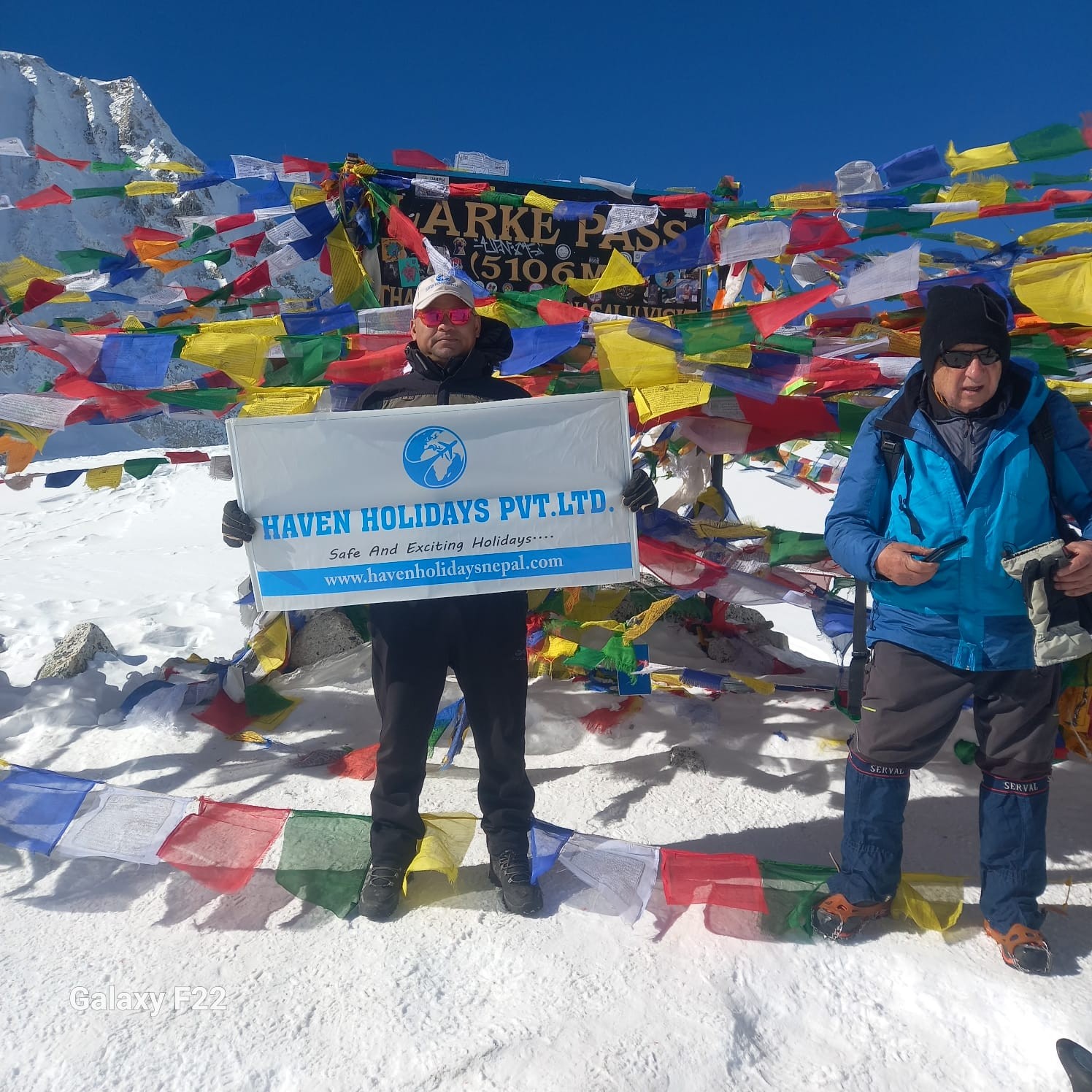 Trekkers crossing Larke Pass surrounded by snow-covered terrain, colorful prayer flags, and panoramic Himalayan peaks on the Manaslu Circuit Trek