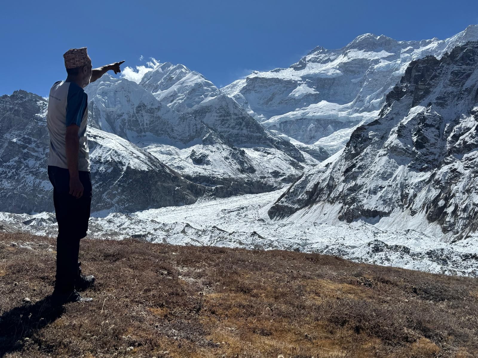Experienced trekking guide standing on the Kanchenjunga Trek trail with Mount Kanchenjunga and surrounding snow-capped Himalayan peaks in eastern Nepal.