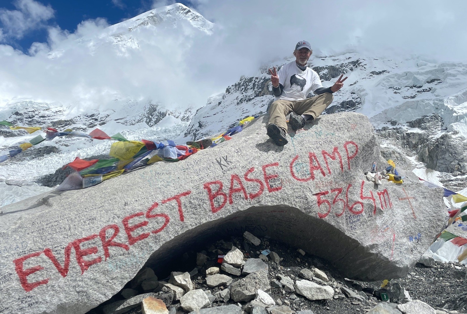 A trekker sits on the iconic Everest Base Camp rock (5,364 m) in September, surrounded by colourful prayer flags, the Khumbu Glacier, fresh snow-covered peaks, and drifting post-monsoon clouds in Nepal's Everest region.