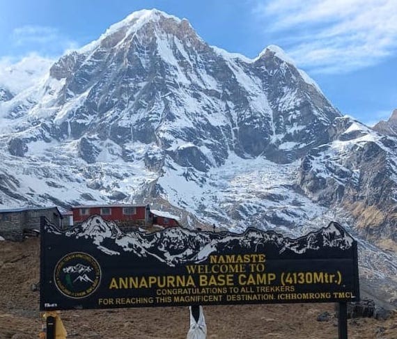 Welcome to Annapurna Base Camp (4,130 m) signboard with the towering Annapurna I massif, glacier, and snow-covered Himalayan peaks in the background under clear blue October skies in Nepal.