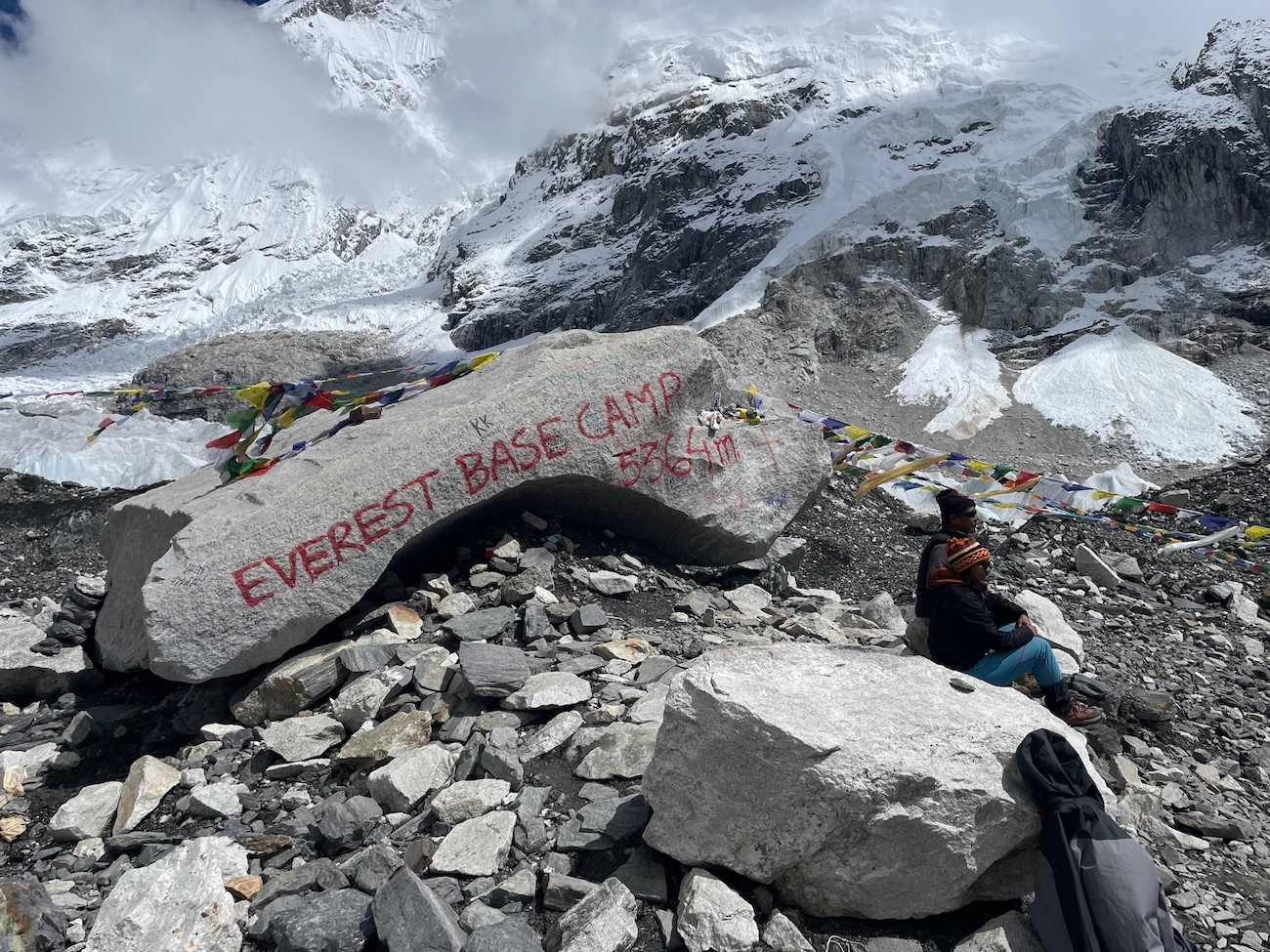 "Trekker standing beside the iconic Everest Base Camp rock with colorful prayer flags, the Khumbu Glacier, and snow-covered Himalayan peaks in the background"  Photo 