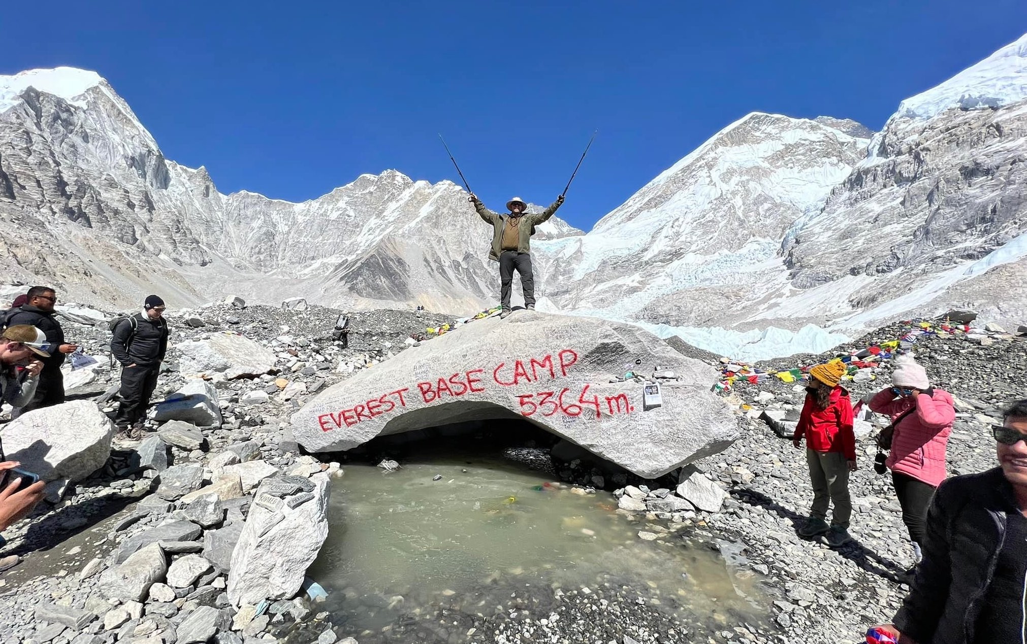 Trekker celebrating at the iconic Everest Base Camp marker (5,364 m), surrounded by the Khumbu Glacier, Mount Everest, Nuptse, and towering Himalayan peaks in Nepal.
