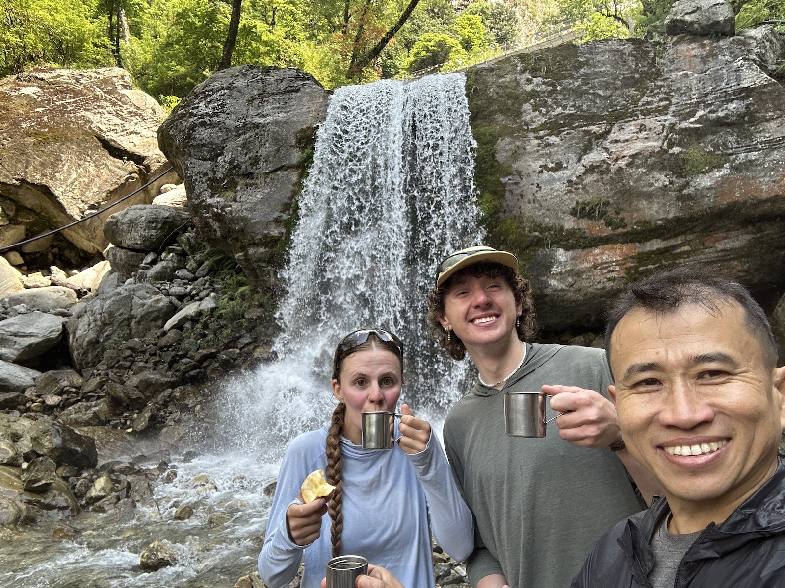 Trekkers enjoying coffee beside a cascading waterfall on the Annapurna Circuit Trek in Nepal, surrounded by lush green forests, rocky cliffs, and Himalayan mountain scenery.