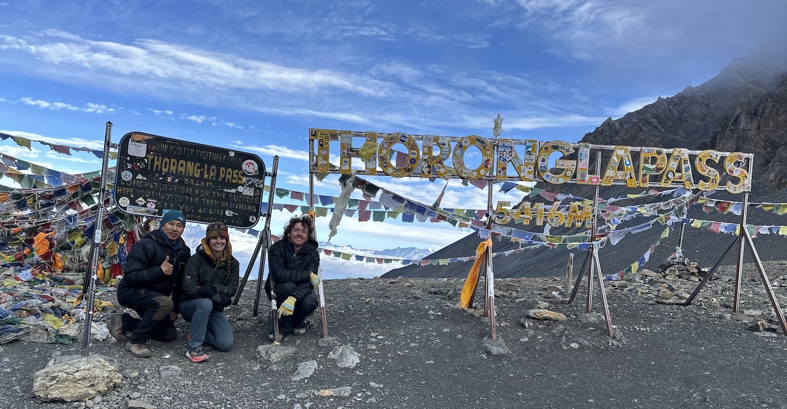 "Thorong La Pass at 5,416 meters on the Annapurna Circuit Trek featuring colorful prayer flags, trekkers, and panoramic views of the snow-covered Himalayan peaks in Nepal."