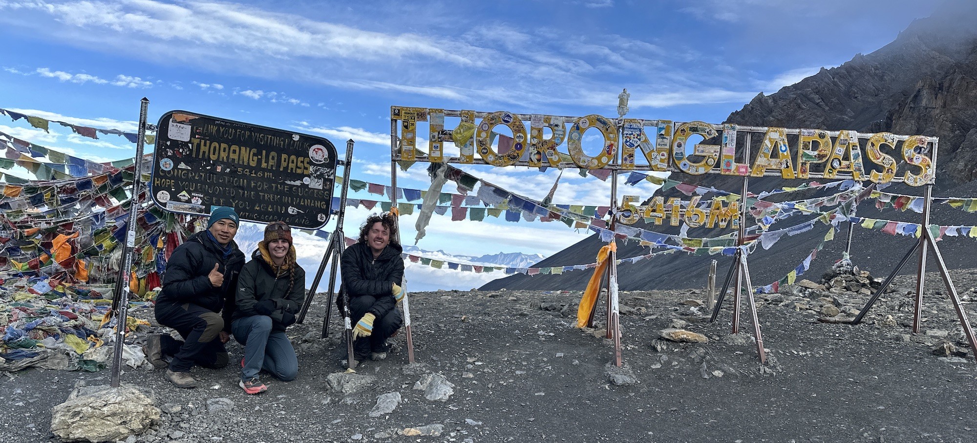 Trekkers pose beside the iconic Thorong La Pass sign at 5,416 metres in September, surrounded by colourful prayer flags, rugged Himalayan landscapes, and clear post-monsoon skies on the Annapurna Circuit Trek.