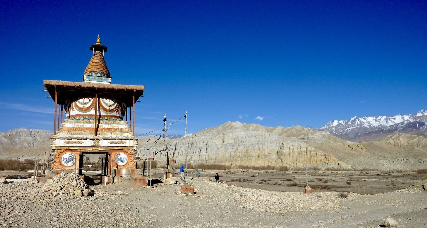 Ancient Buddhist stupa in Upper Mustang, Nepal, decorated with colorful prayer flags and set against dramatic desert cliffs and snow-capped Himalayan peaks.