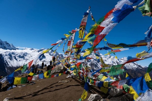 Trekkers hiking toward Kyanjin Ri through rocky alpine terrain with colorful prayer flags and snow-capped Himalayan peaks in the background.